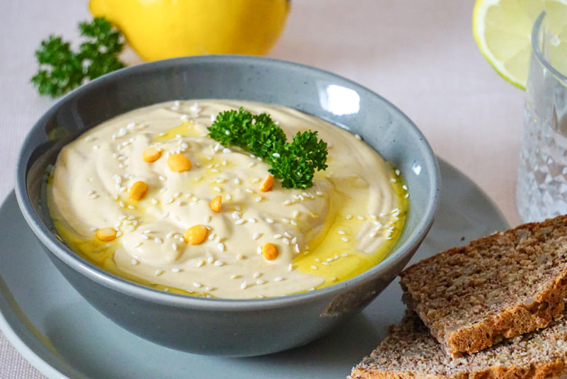 Eine Schale mit Hummus, zubereitet nach dem Hummus Rezept, steht auf dem Tisch, daneben liegt frisches Brot