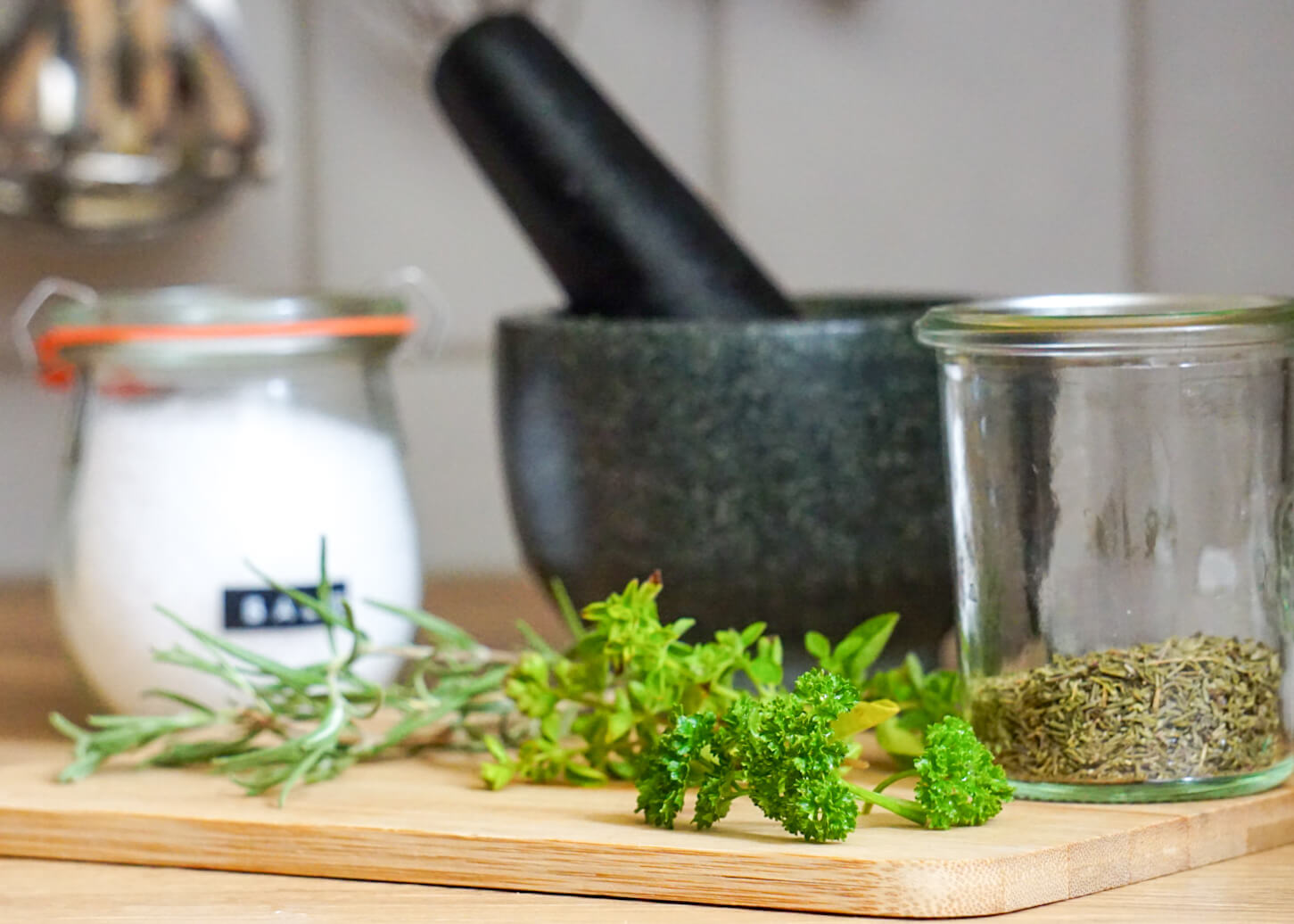 Frische Kräuter (Rosmarin, Oregano, Pertersilie und Thymian) liegen auf Holzbrettchen. Dahinter steht ein Mörser