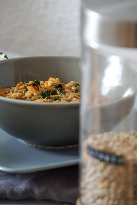 Eine Schale mit Kokos-Linsen-Curry, dazu Reis im Hintergrund, Vordergrund zeigt unscharf ein Glas mit Linsen