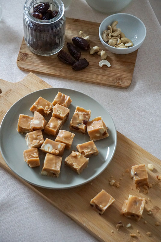 Kleine Stücle Karamell-Fudge auf Holzbrett, dazu Datteln und Cashewkerne