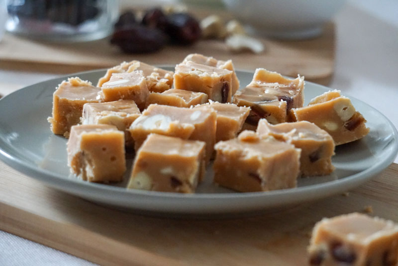 Kleine Stücle Karamell-Fudge auf Holzbrett, dazu Datteln und Cashewkerne